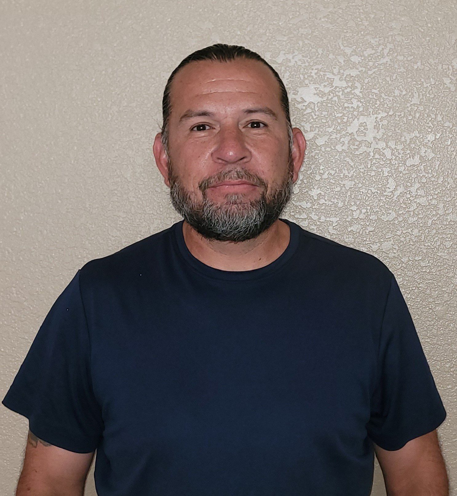Smiling man with a beard wearing a navy blue t-shirt against a plain wall.