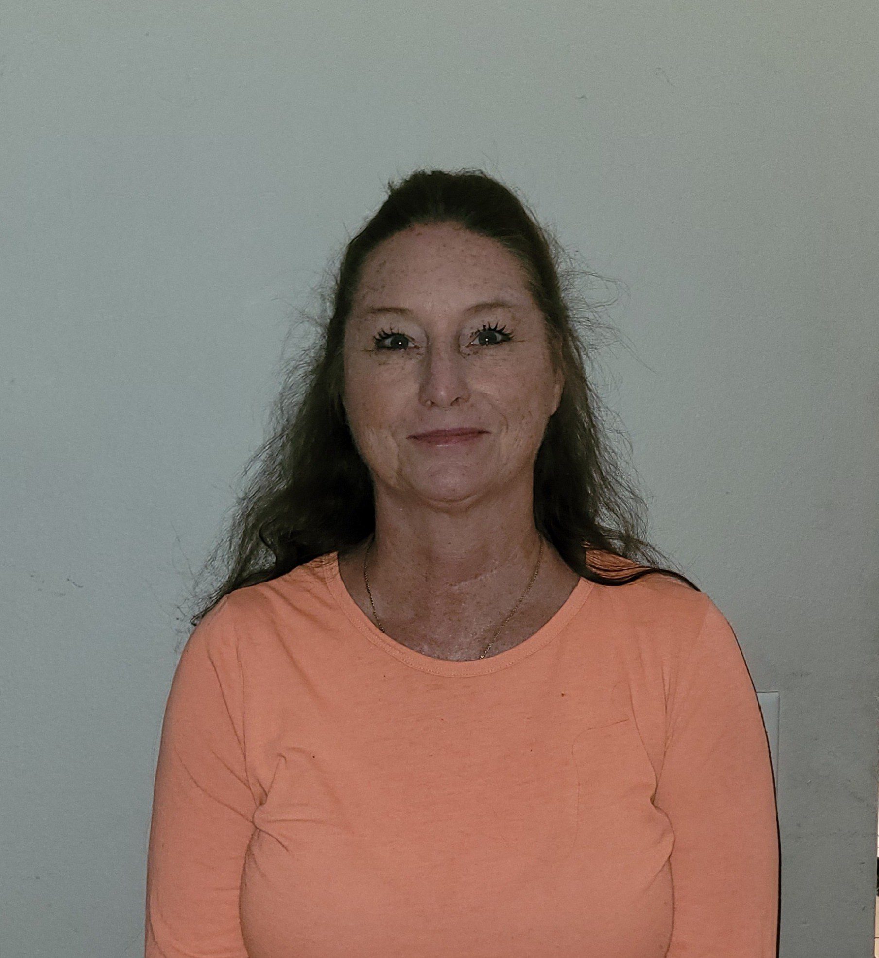 A woman with long dark hair wearing an orange shirt against a plain background.