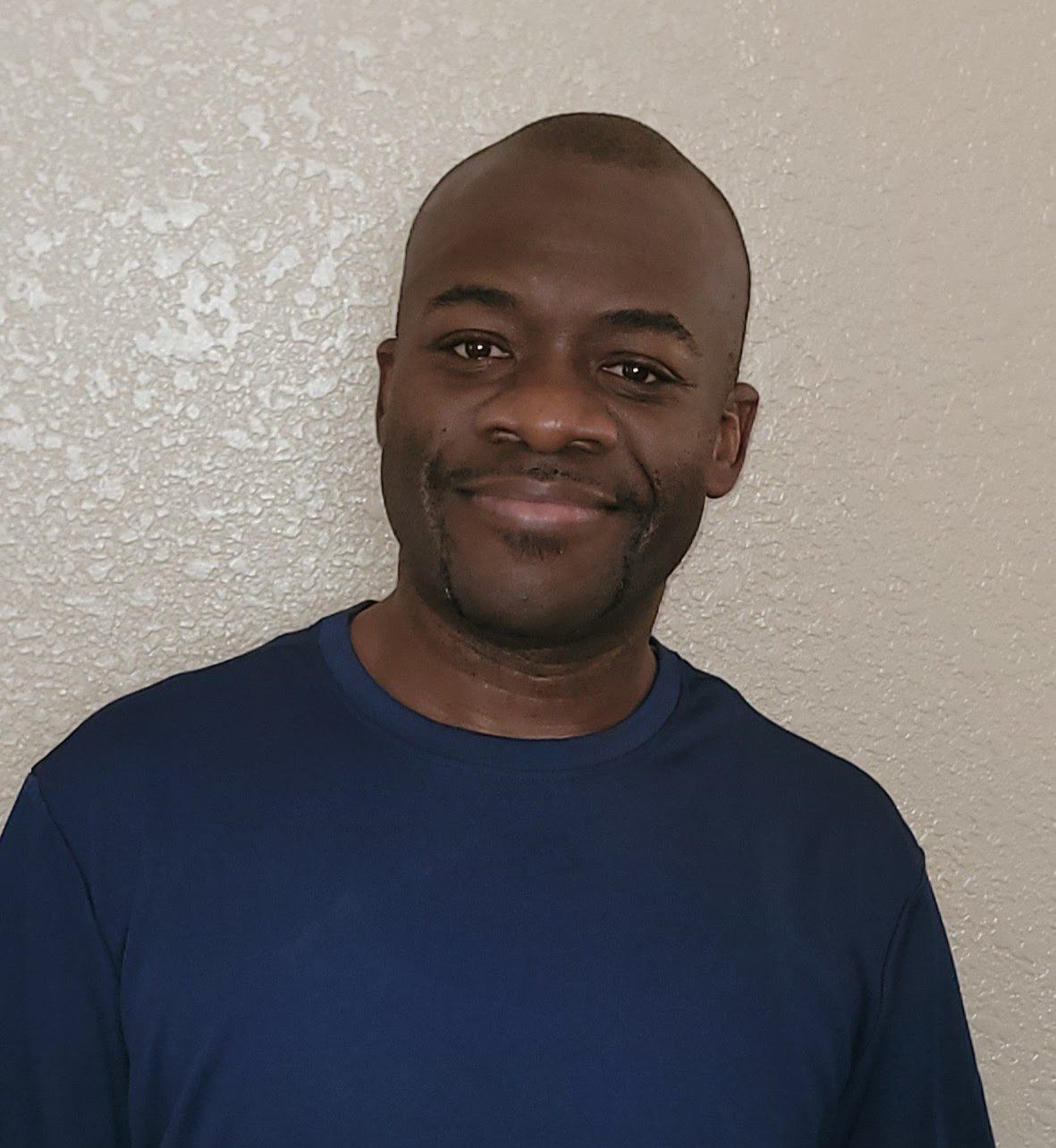 Smiling man in a navy blue shirt against a beige wall.